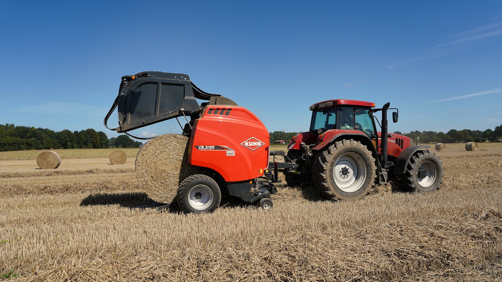 Tractor pulling a round baler harvesting hay bales in a field at sunset